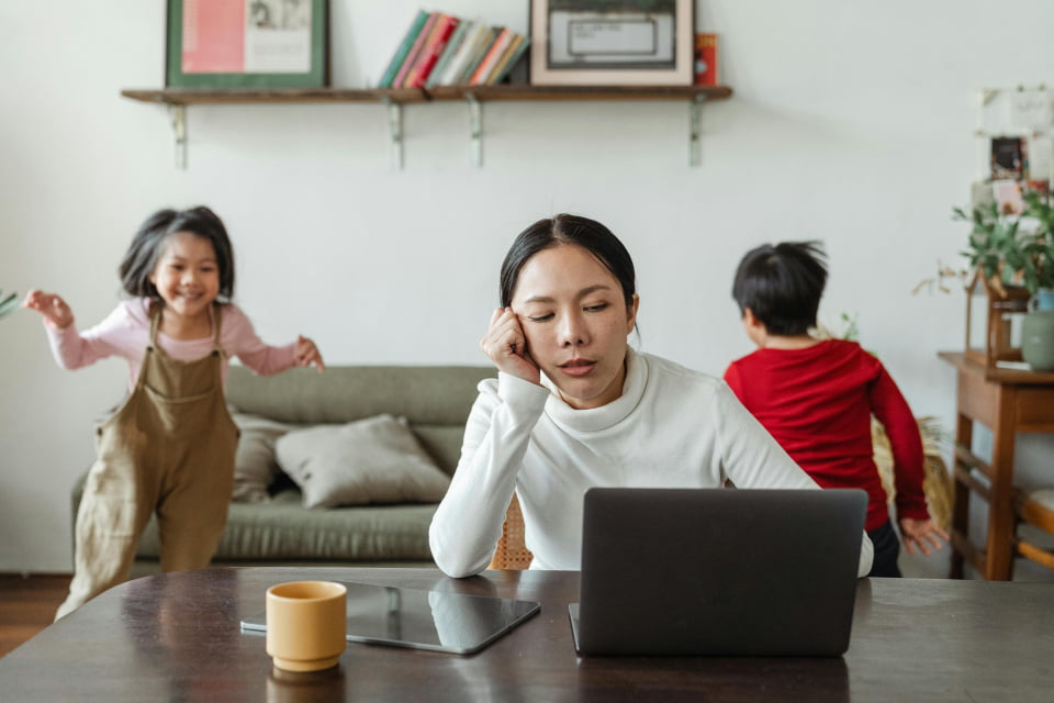 tired woman getting remote counseling looking at laptop with kids running around sm